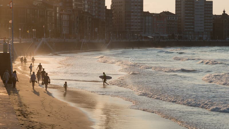 San Lorenzo, Gijon beach and waterfront, at sunset. Unkown people on the sand; a surfer coming out of the sea with his board. Asturias, Spain, Europe