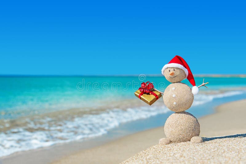 Sandig snögubbe för Smiley på stranden i julhatt med den guld- gåvan