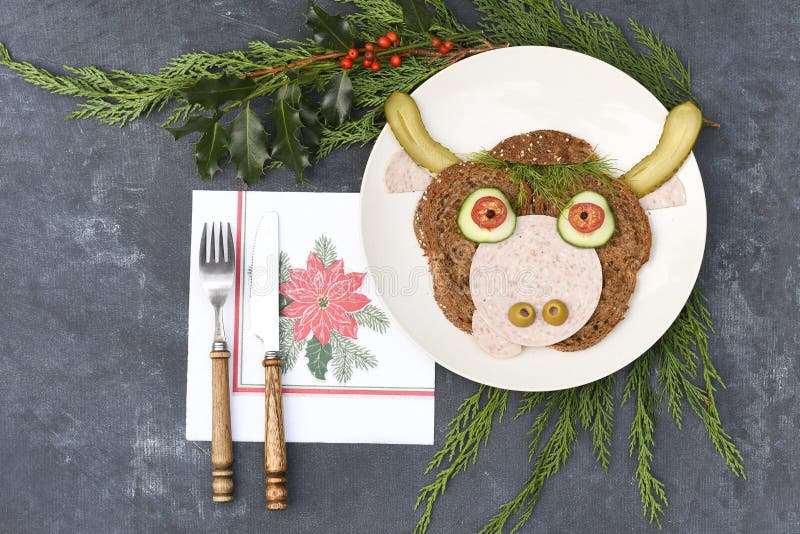 Sandwich in the form of a portrait of a cheerful bull made of dark bread, sausages and vegetables on a plate, on a dark background with a decor of thuja and holly branches. Sandwich in the form of a portrait of a cheerful bull made of dark bread, sausages and vegetables on a plate, on a dark background with a decor of thuja and holly branches