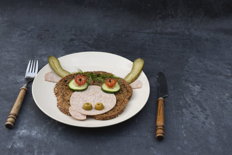 Sandwich in the form of a portrait of a cheerful bull made of dark bread, sausages and vegetables on a plate, on a dark background with a decor of thuja and holly branches. High quality photo. Sandwich in the form of a portrait of a cheerful bull made of dark bread, sausages and vegetables on a plate, on a dark background with a decor of thuja and holly branches. High quality photo