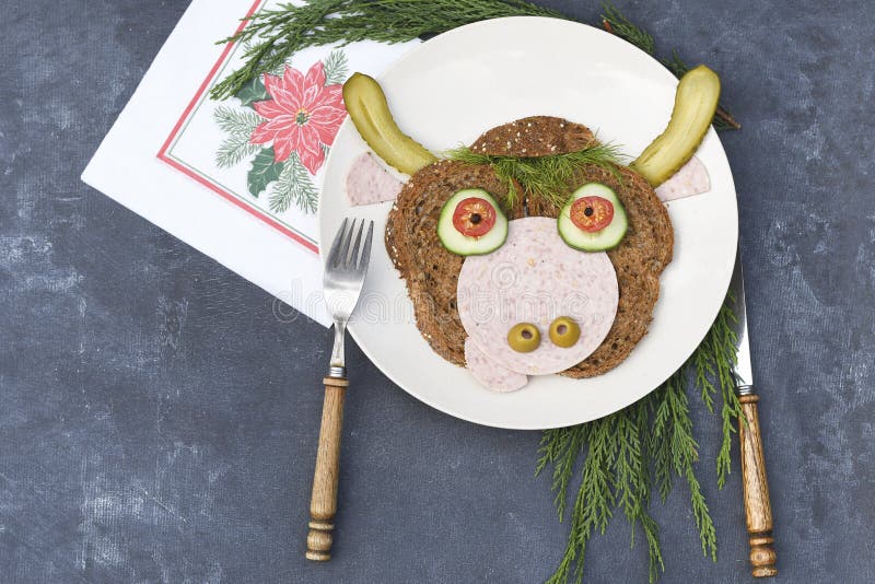 Sandwich in the form of a portrait of a cheerful bull made of dark bread, sausages and vegetables on a plate, on a dark background with a decor of thuja and holly branches. Sandwich in the form of a portrait of a cheerful bull made of dark bread, sausages and vegetables on a plate, on a dark background with a decor of thuja and holly branches