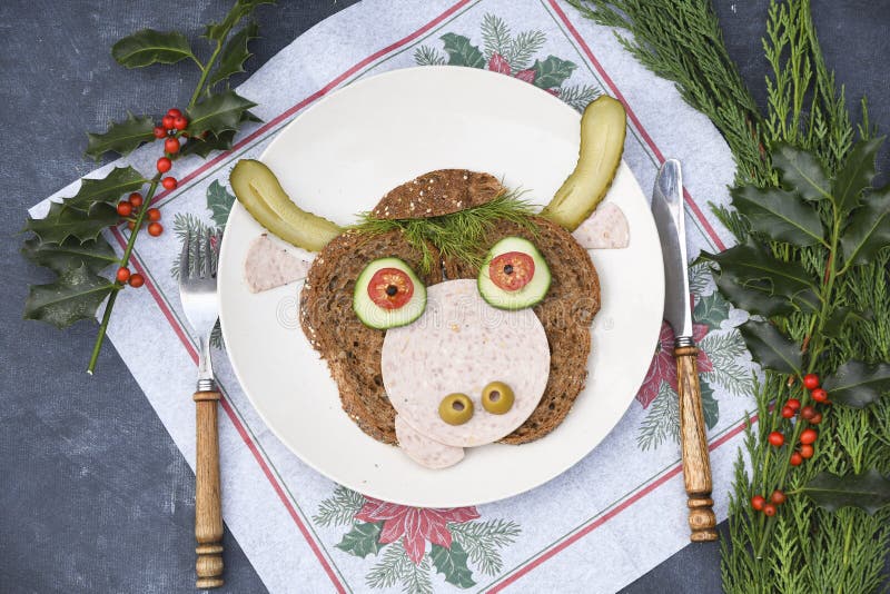 Sandwich in the form of a portrait of a cheerful bull made of dark bread, sausages and vegetables on a plate, on a dark background with a decor of thuja and holly branches. Sandwich in the form of a portrait of a cheerful bull made of dark bread, sausages and vegetables on a plate, on a dark background with a decor of thuja and holly branches