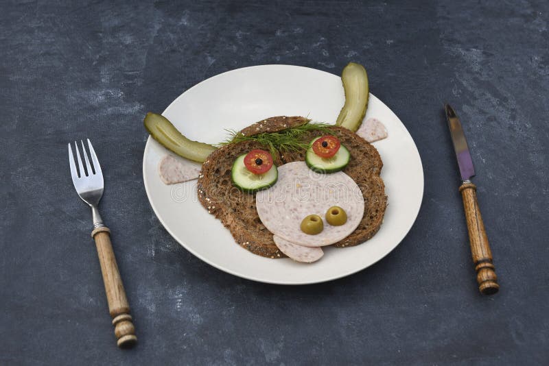 Sandwich in the form of a portrait of a cheerful bull made of dark bread, sausages and vegetables on a plate, on a dark background with a decor of thuja and holly branches. High quality photo. Sandwich in the form of a portrait of a cheerful bull made of dark bread, sausages and vegetables on a plate, on a dark background with a decor of thuja and holly branches. High quality photo