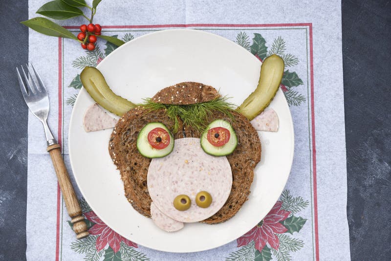 Sandwich in the form of a portrait of a cheerful bull made of dark bread, sausages and vegetables on a plate, on a dark background with a decor of thuja and holly branches. Sandwich in the form of a portrait of a cheerful bull made of dark bread, sausages and vegetables on a plate, on a dark background with a decor of thuja and holly branches