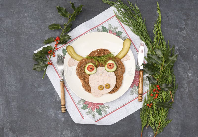 Sandwich in the form of a portrait of a cheerful bull made of dark bread, sausages and vegetables on a plate, on a dark background with a decor of thuja and holly branches. Sandwich in the form of a portrait of a cheerful bull made of dark bread, sausages and vegetables on a plate, on a dark background with a decor of thuja and holly branches