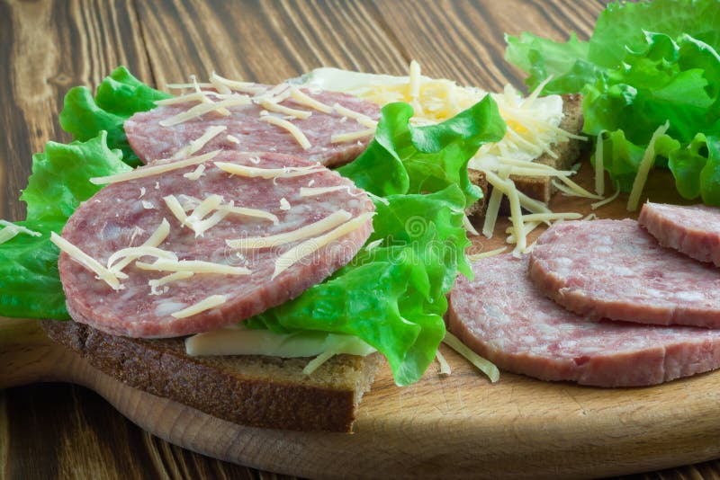 Sandwichs des saucisses de pain et de beurre, de laitue et de salami arrosées avec du fromage râpé sur une planche à découper et photos stock Sandwichs des saucisses de pain et de beurre, de laitue et de salami arrosées avec du fromage râpé sur une planche à découper et photos stock