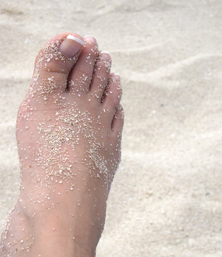 A lounging woman's French pedicured right foot lightly dusted with white beach sand, above the sandy beach in the background. A lounging woman's French pedicured right foot lightly dusted with white beach sand, above the sandy beach in the background
