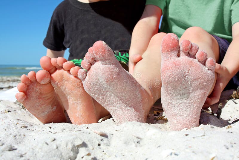 Two young children's feet are seen, covered in sand as they sit on the beach by the ocean. Two young children's feet are seen, covered in sand as they sit on the beach by the ocean