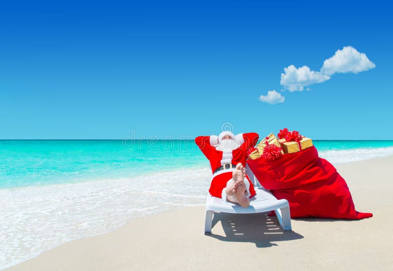 Santa Claus with Christmas sack full of gifts relax on sunlounger barefooted at perfect sandy ocean beach.