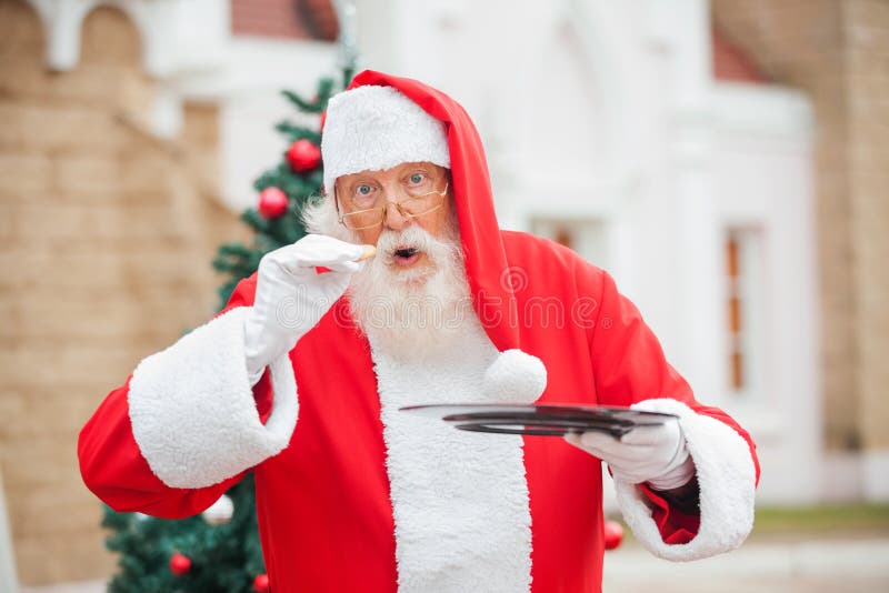 Portrait of senior man dressed as Santa Claus eating cookie against house