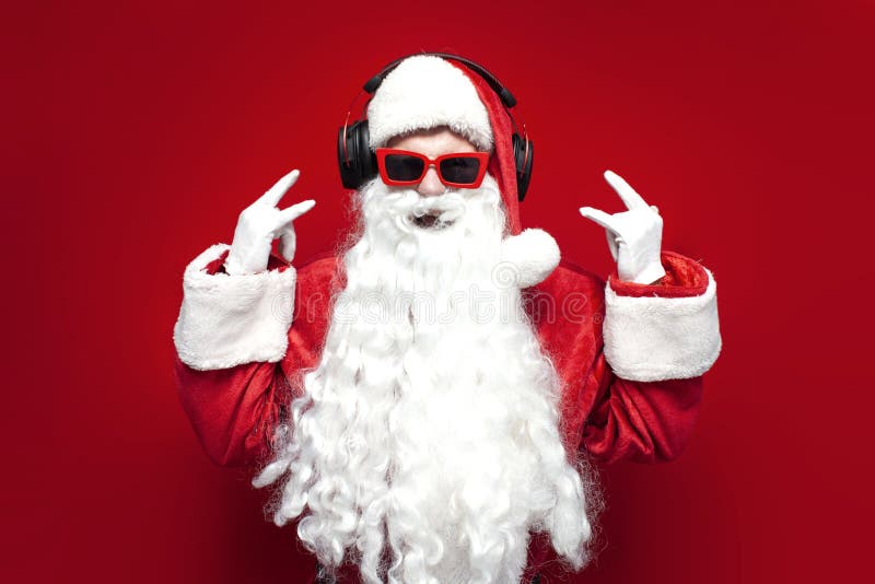 Santa claus en chapeau et lunettes festives écoute de la musique dans les casques et chante sur fond rouge images stock