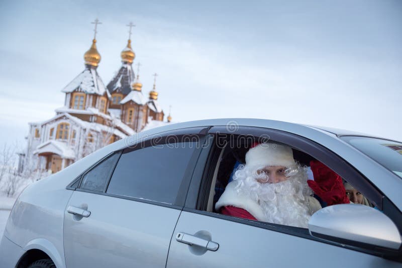 Anadyr, Chukotka, Russia - December 31, 2013. Santa Claus and Snow Maiden in the car against the background of the winter church. Father Christmas waves his hand in greeting. New Year's Eve Day. Anadyr, Chukotka, Russia - December 31, 2013. Santa Claus and Snow Maiden in the car against the background of the winter church. Father Christmas waves his hand in greeting. New Year's Eve Day