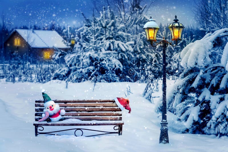 Santa hat and New Year`s soft toy on a bench in the winter forest against the background of a village house and lantern. Christma