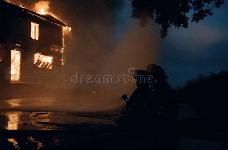 Home fully ablaze with flames leaping from roof, windows and silhouetted firefighters aiming firehose at flaming home. Home fully ablaze with flames leaping from roof, windows and silhouetted firefighters aiming firehose at flaming home.