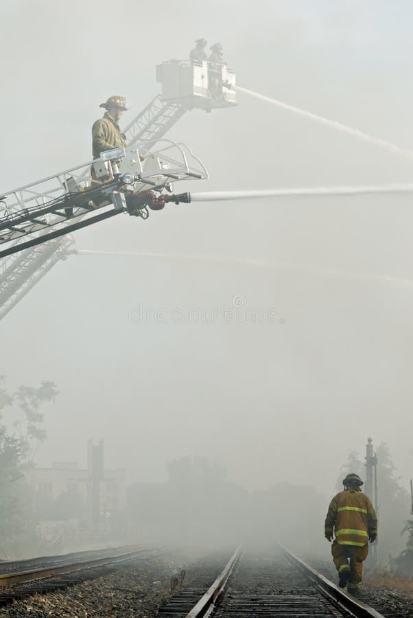Firefighters shoot water through smoke across railroad track while another fireman walks down the track. Firefighters shoot water through smoke across railroad track while another fireman walks down the track.