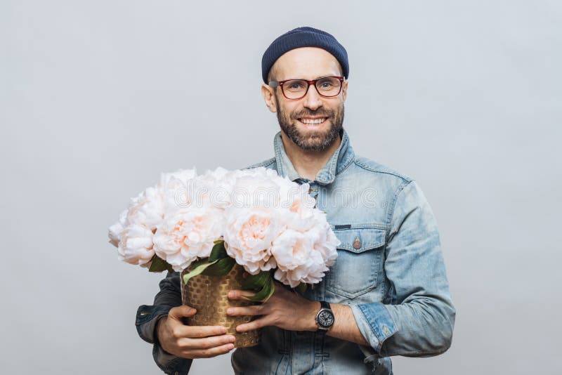 Satisfied bearded male with cheerful expression has broad smile, holds bunch of flowers, wears hat and denim jacket, isolated stock images Satisfied bearded male with cheerful expression has broad smile, holds bunch of flowers, wears hat and denim jacket, isolated stock images