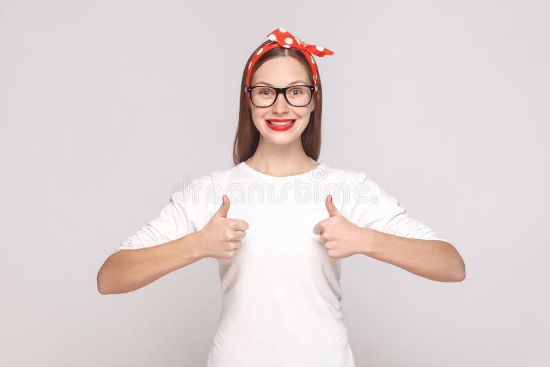 I am satisfied, thumbs up. portrait of beautiful emotional young woman in white t-shirt with freckles, black glasses, red lips and head band. indoor studio shot, on light gray background. I am satisfied, thumbs up. portrait of beautiful emotional young woman in white t-shirt with freckles, black glasses, red lips and head band. indoor studio shot, on light gray background.