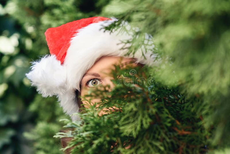 Scared, open-eyed look of a beautiful girl in a santa claus hat hiding behind a christmas tree. Concept.