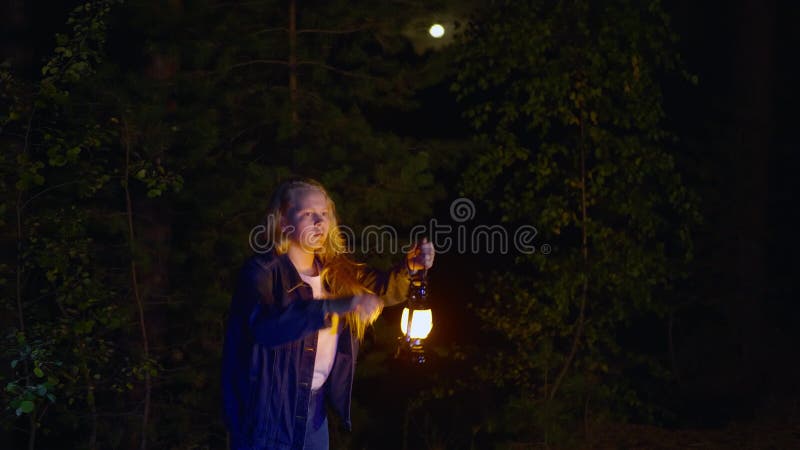 Scared young girl holding oil lamp and looking at something in dark forest at night