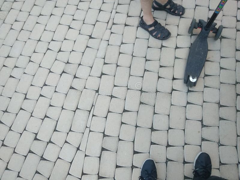 Scarpe dei piedi delle persone che stanno sul ciottolo grigio che pavimenta su una via e su un trotinet fotografia stock libera da diritti Scarpe dei piedi delle persone che stanno sul ciottolo grigio che pavimenta su una via e su un trotinet fotografia stock libera da diritti