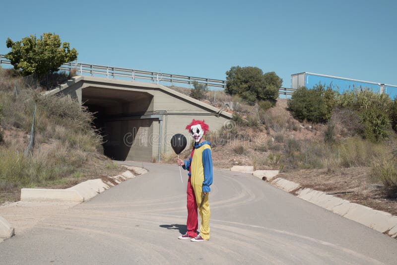 A scary clown wearing a colorful yellow, red and blue costume outdoors, holding a black balloon in his hand, standing in the middle of a secondary road, at the entrance of a tunnel. A scary clown wearing a colorful yellow, red and blue costume outdoors, holding a black balloon in his hand, standing in the middle of a secondary road, at the entrance of a tunnel