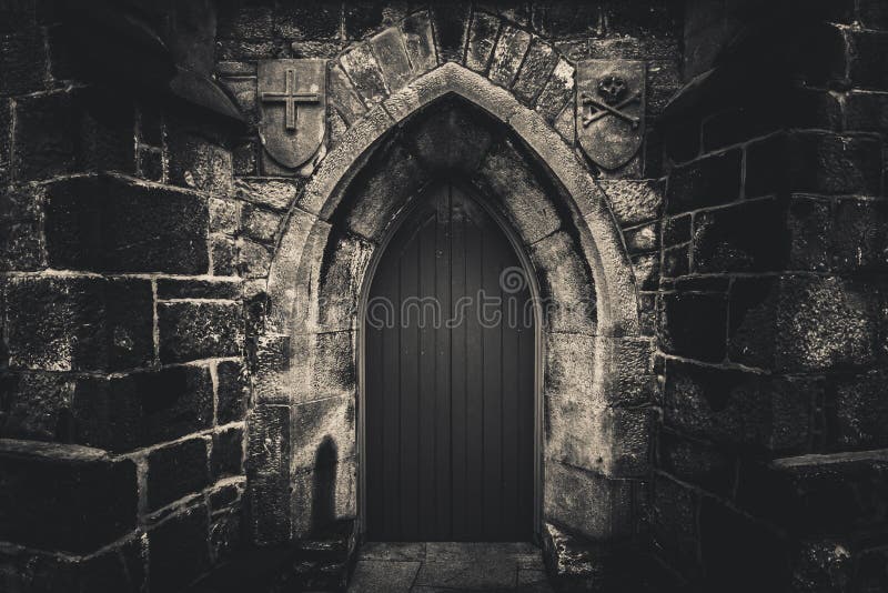 Scary pointy wooden door in an old and wet stone wall building with cross, skull and bones at both sides in black and white. Concept mystery, death and danger, architecture, church, dark, entrance, light, spooky, castle, ancient, architectural, brick, cathedral, gothic, moody, religion, ruin, style, rust, horror, christ, spiritual, traditional, bible, god, holy, vintage, halloween, haunted, torn, shape, texture, creepy, alone