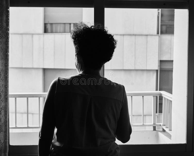 Scatto in scala di grigi sul retro di una donna all'interno della casa mentre guarda fuori dalla finestra fotografia stock