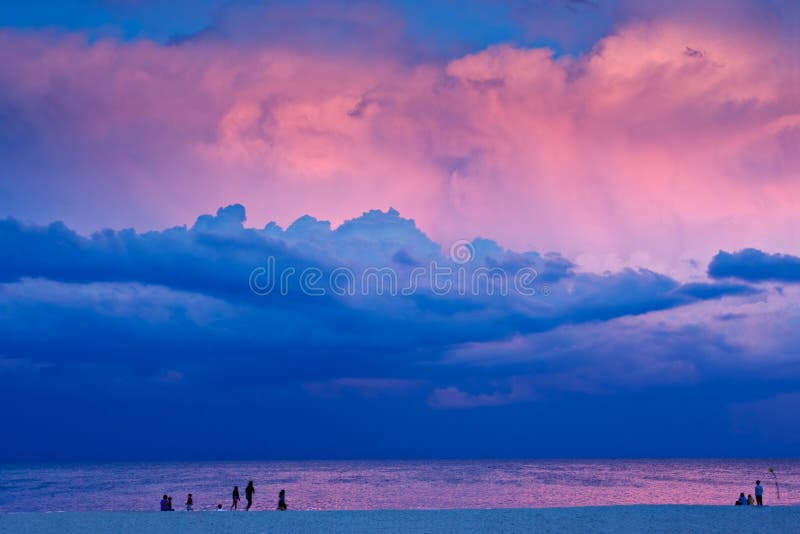 A beach scene in the evening at sunset. There are people on the beach with a dramatic stormy sky overhead. The last rays of sunlight are lighting some of the clouds. A beach scene in the evening at sunset. There are people on the beach with a dramatic stormy sky overhead. The last rays of sunlight are lighting some of the clouds.
