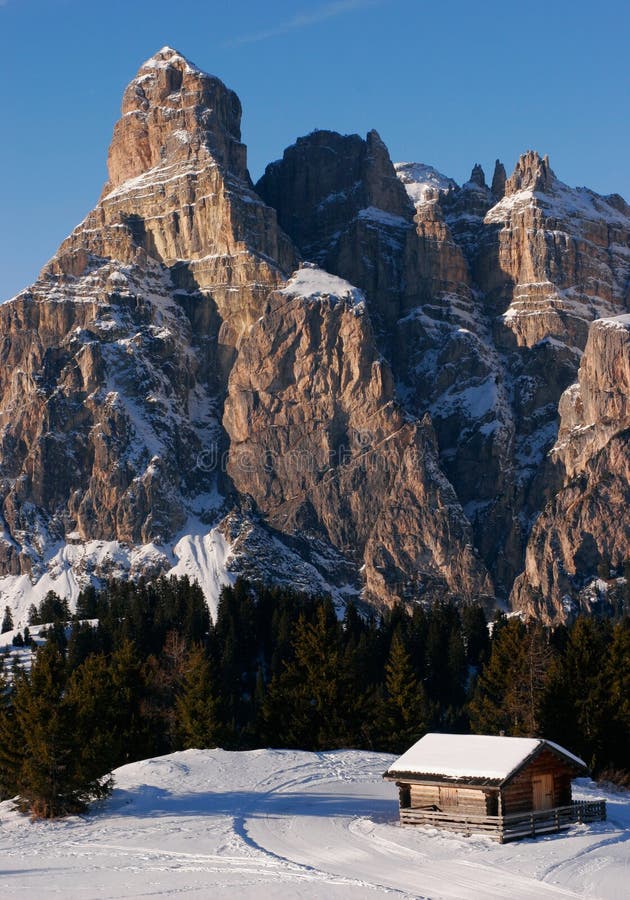 Small wooden mountain cabin with a very impressive rock formation in the background. Taken in the vicinity of the village of Corvara in the Dolomites, Alta Badia, Alto Adige, Italy. The late afternoon sun casts soft shadows beautifully illuminates the rocks, trees and cabin. Skiing tracks are in the foreground. Small wooden mountain cabin with a very impressive rock formation in the background. Taken in the vicinity of the village of Corvara in the Dolomites, Alta Badia, Alto Adige, Italy. The late afternoon sun casts soft shadows beautifully illuminates the rocks, trees and cabin. Skiing tracks are in the foreground.