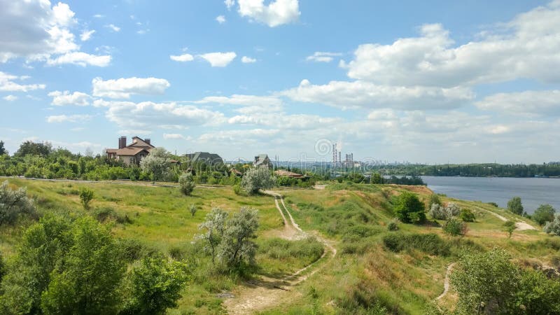 Sceniskt landskap som förbiser kullen med banor, en flod, landshus, blå molnig himmel och röka rör arkivfoto Sceniskt landskap som förbiser kullen med banor, en flod, landshus, blå molnig himmel och röka rör arkivfoto