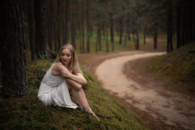 Sch?ne junge Blondine, die in der Waldnymphe im wei?en Kleid im immergr?nen Holz sitzen lizenzfreie stockfotos Sch?ne junge Blondine, die in der Waldnymphe im wei?en Kleid im immergr?nen Holz sitzen lizenzfreie stockfotos