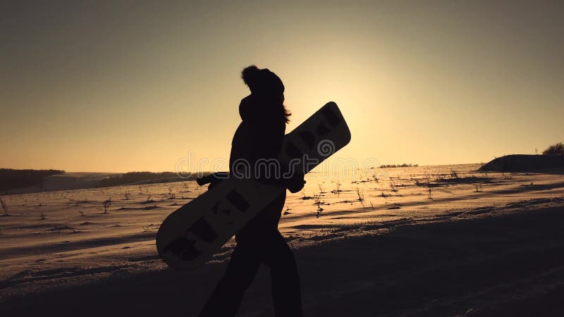 Schattenbildmädchen-Snowboarderweg mit Snowboard auf die Gebirgsoberseite bei Sonnenuntergang