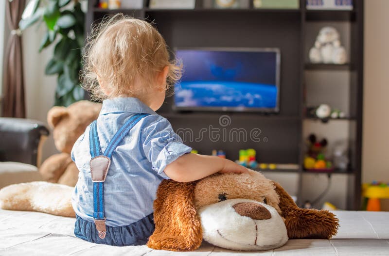 Cute baby boy and his dog plush toy watching TV sitting on a couch in the living room at home. Cute baby boy and his dog plush toy watching TV sitting on a couch in the living room at home.