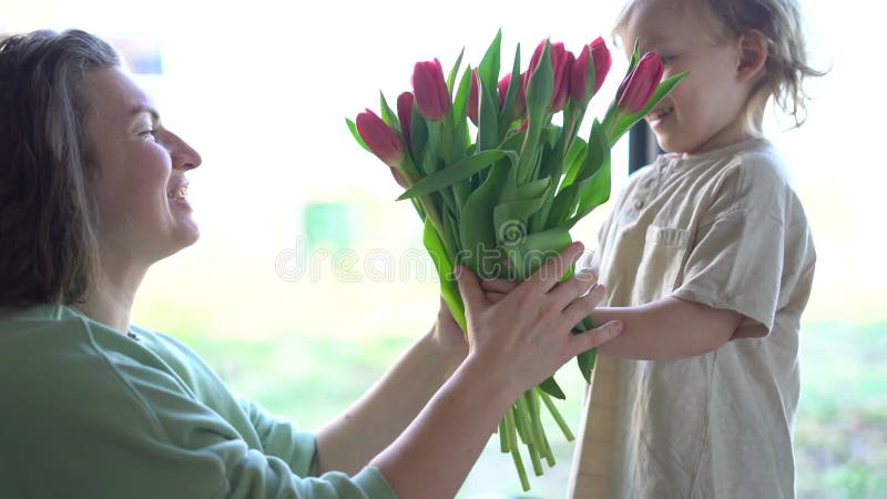 Schattige blond jongen geeft zijn moeder een mooie verse bouquet rode tulpen. moeder - en zoon - moeders dag