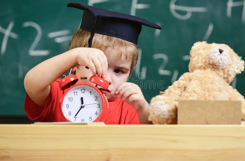 Scherzi il ragazzo in cappuccio accademico vicino al microscopio, tenute cronometrano in aula, lavagna su fondo Il bambino sul fr immagine stock Scherzi il ragazzo in cappuccio accademico vicino al microscopio, tenute cronometrano in aula, lavagna su fondo Il bambino sul fr immagine stock