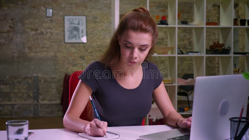 Schieten van werkend Kaukasisch roodharig wijfje, geconcentreerd op haar het werk, zittings en het maken nota's, het controleren