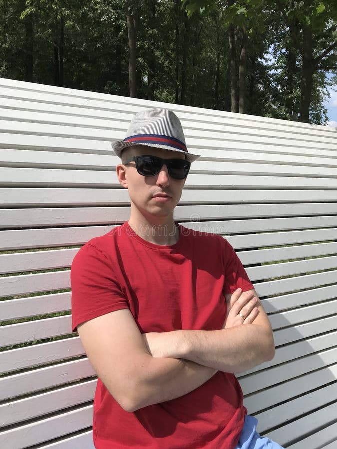 Schließen Sie oben vom kaukasischen jungen Mann im Hut, sitzt rotes T-Shirt und Sonnenbrille auf einer weißen Bank im Park und Bl lizenzfreie stockfotografie Schließen Sie oben vom kaukasischen jungen Mann im Hut, sitzt rotes T-Shirt und Sonnenbrille auf einer weißen Bank im Park und Bl lizenzfreie stockfotografie
