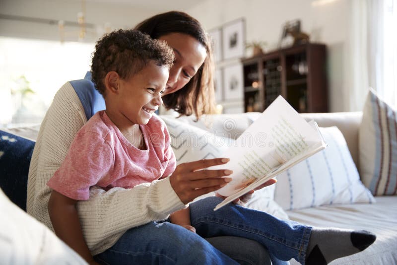 Schließen Sie oben von der jungen Mutter, die auf einem Sofa im Wohnzimmer sitzt, das ein Buch mit ihrem Kleinkindsohn liest, der