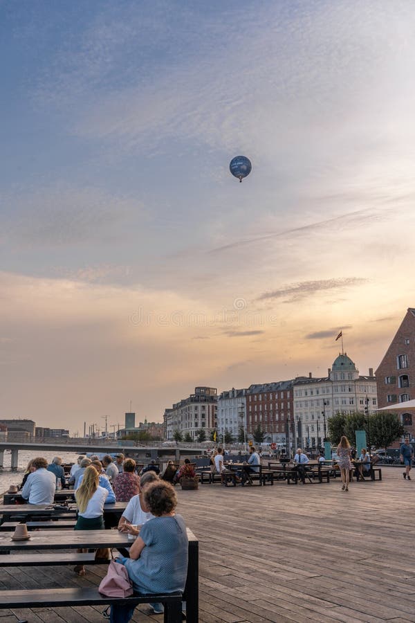 People sitting at the tables of open-air dining area of Café og Restaurant Ofelia in Nyhavn - Københavns Havn - the central tourist area of Copenhagen, Denmark, on a warm evening, at sunset, with an aerostat - hot air baloon - flying in the sky above. People sitting at the tables of open-air dining area of Café og Restaurant Ofelia in Nyhavn - Københavns Havn - the central tourist area of Copenhagen, Denmark, on a warm evening, at sunset, with an aerostat - hot air baloon - flying in the sky above