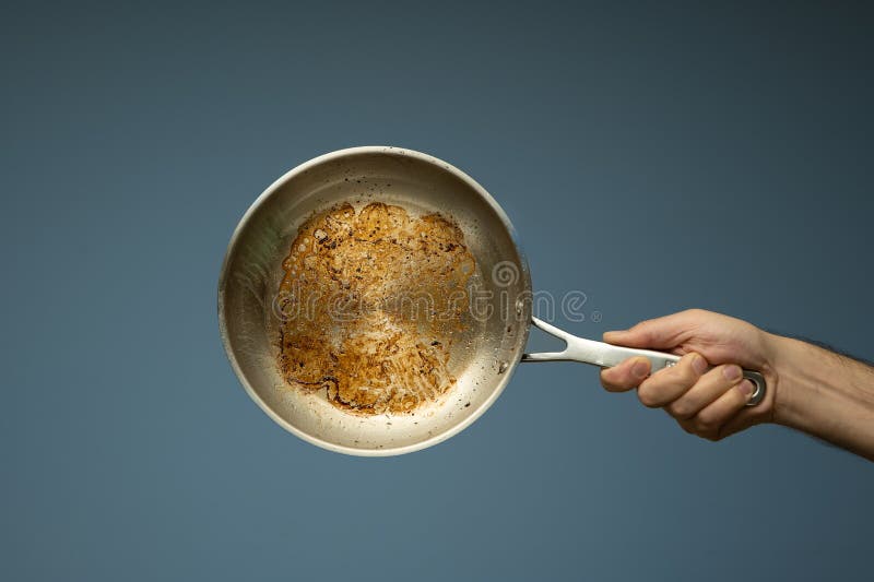 Dirty oily burnt metal frying pan held in hand by a male hand. Close up studio shot, isolated on a light blue background. Dirty oily burnt metal frying pan held in hand by a male hand. Close up studio shot, isolated on a light blue background.