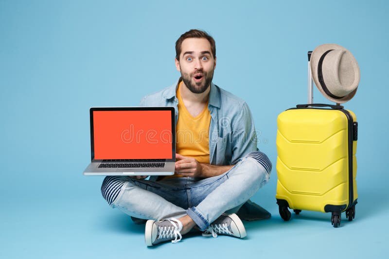 Shocked traveler tourist man in yellow clothes isolated on blue background. Passenger traveling abroad on weekend. Air flight journey concept. Sit near suitcase, hold laptop with blank empty screen. Shocked traveler tourist man in yellow clothes isolated on blue background. Passenger traveling abroad on weekend. Air flight journey concept. Sit near suitcase, hold laptop with blank empty screen