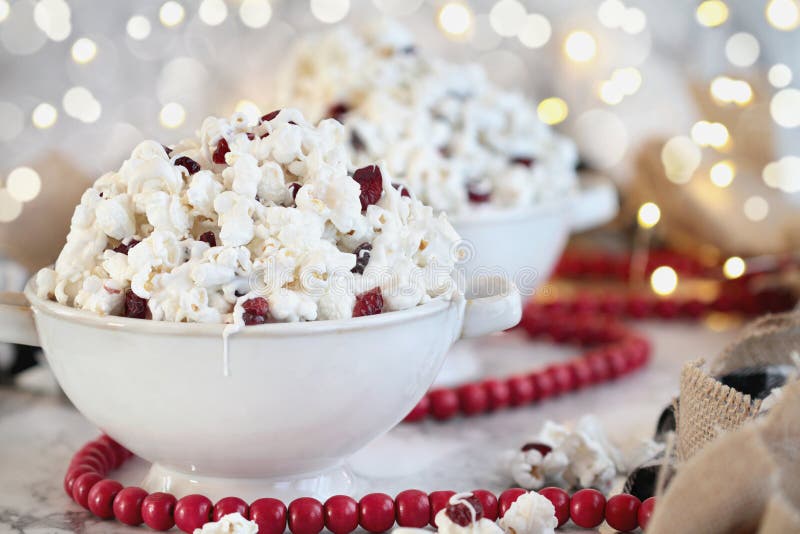 Bowls of homemade popcorn and dried cranberry snack covered in white chocolate ready for the holidays surrounded by bokeh lights and red bead garland.. Selective focus with blurred background. Bowls of homemade popcorn and dried cranberry snack covered in white chocolate ready for the holidays surrounded by bokeh lights and red bead garland.. Selective focus with blurred background
