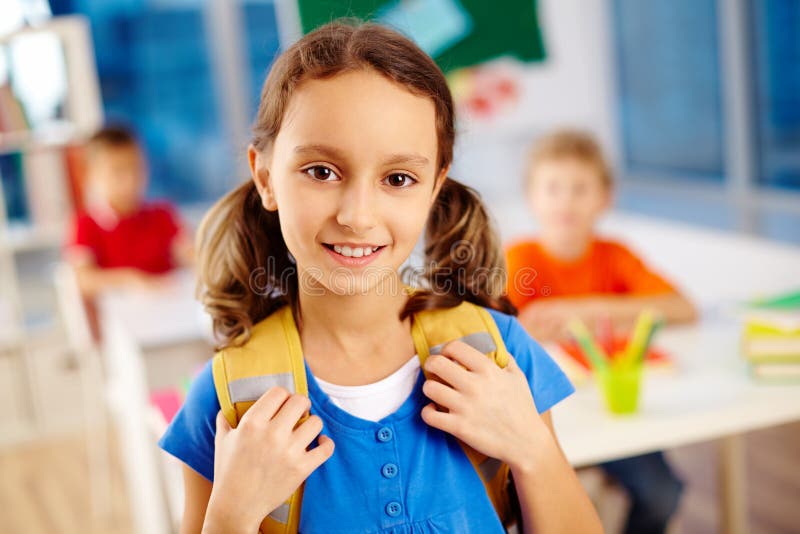 Portrait of cute schoolgirl with backpack looking at camera with classmates on background