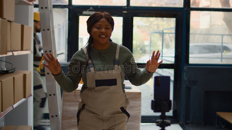 Warehouse chief supply chain officer using smartphone to make training video for interns. African american supervisor films herself in depository showing trainees how to seal cardboard boxes. Warehouse chief supply chain officer using smartphone to make training video for interns. African american supervisor films herself in depository showing trainees how to seal cardboard boxes