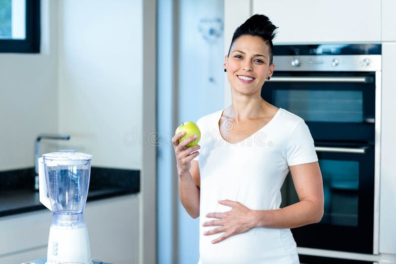 Pregnant woman holding a green apple and smiling in kitchen. Pregnant woman holding a green apple and smiling in kitchen