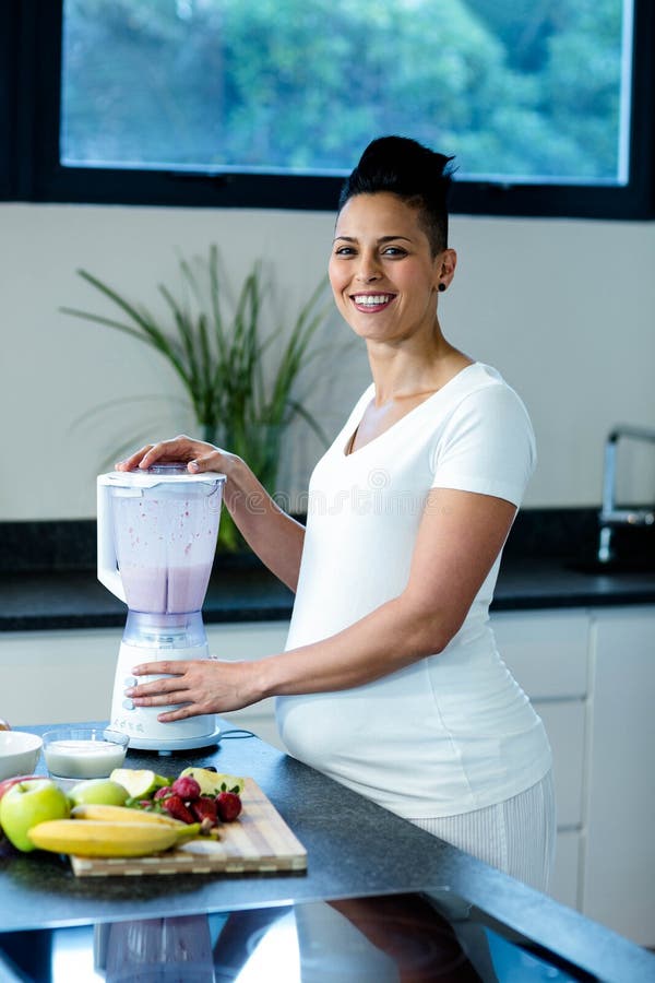 Pregnant woman standing in kitchen and preparing fruit juice in blender. Pregnant woman standing in kitchen and preparing fruit juice in blender