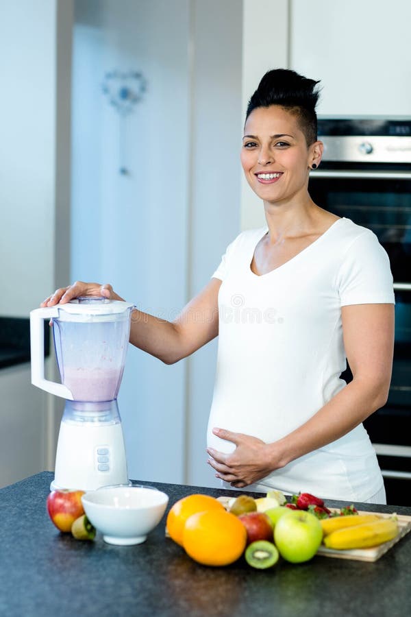 Pregnant woman standing with hand on belly while preparing fruit juice in blender. Pregnant woman standing with hand on belly while preparing fruit juice in blender