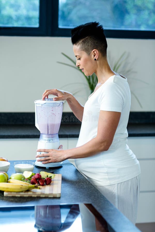 Pregnant woman standing in kitchen and preparing fruit juice in blender. Pregnant woman standing in kitchen and preparing fruit juice in blender