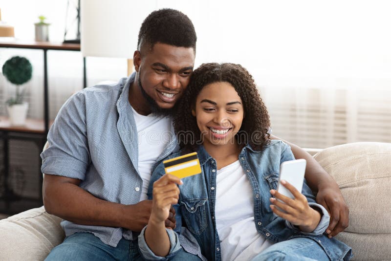 Happy black couple buying goods online, using smartphone and credit card at home, enjoying shopping in internet, free space. Happy black couple buying goods online, using smartphone and credit card at home, enjoying shopping in internet, free space