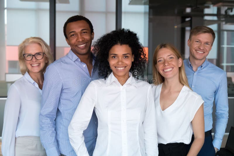 Attractive successful people standing together smiling posing looking at camera feels proud and satisfied. Multi-ethnic businesspeople led by afro mixed race women photographing in modern office room. Attractive successful people standing together smiling posing looking at camera feels proud and satisfied. Multi-ethnic businesspeople led by afro mixed race women photographing in modern office room